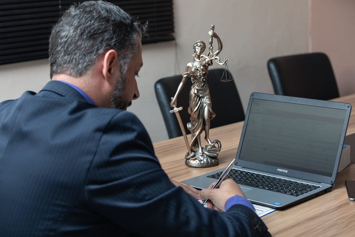 Symbolbild: Bronze-Statue der Justitia mit Waage und Schwert auf einem Tisch neben einem Laptop