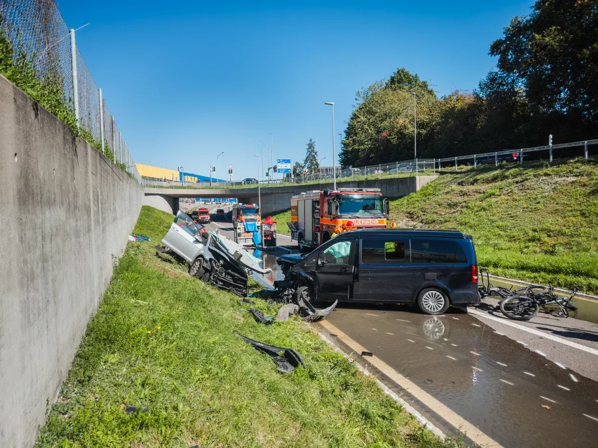 Ein schwarzer Van und ein heller Pkw sind auf einer Autobahnausfahrt kollidiert. Feuerwehrfahrzeuge sind vor Ort.