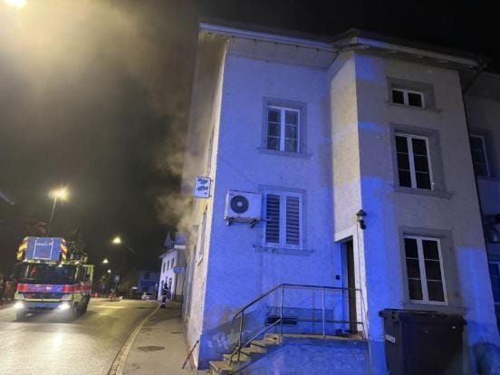 Rechts ein Gebäude, aus dem Rauch aus den Fenstern kommt. Links davon ein Feuerwehrauto.