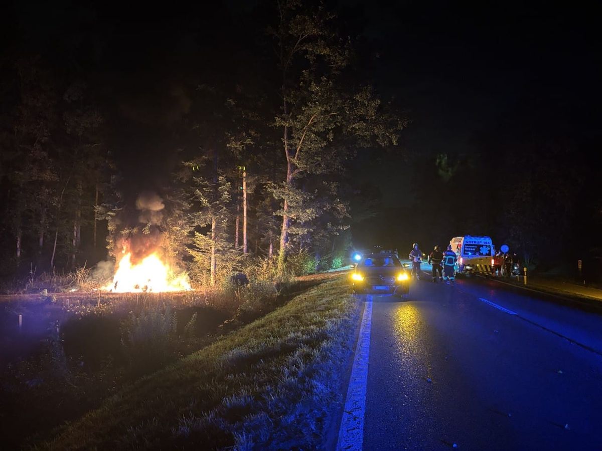Auto in Vollbrand am Waldrand und auf der Strasse ein Rettungswagen und Feuerwehrleute.