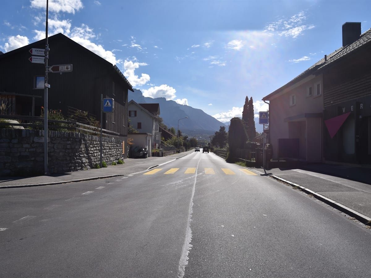 Blick auf den Fussgängerstreifen. Links und rechts der Strasse sind Häuser. Grösstenteils heiterer Himmel.