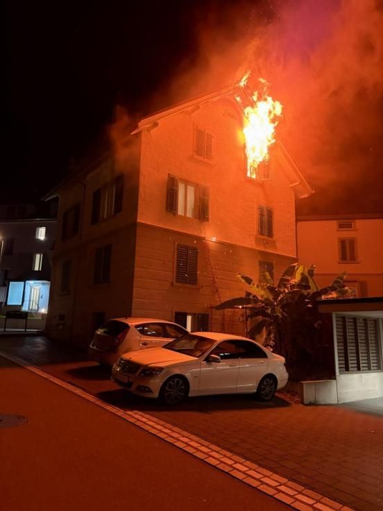 Aus einem Fenster eines Mehrfamilienhauses steigen Flammen empor. Es ist Nacht.