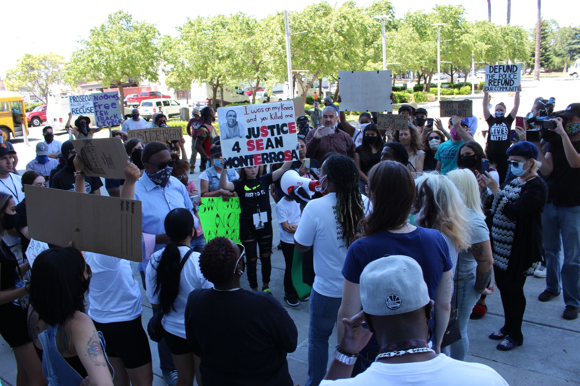 A group of about 50 protesters holding signs in a plaza surrounded by trees.