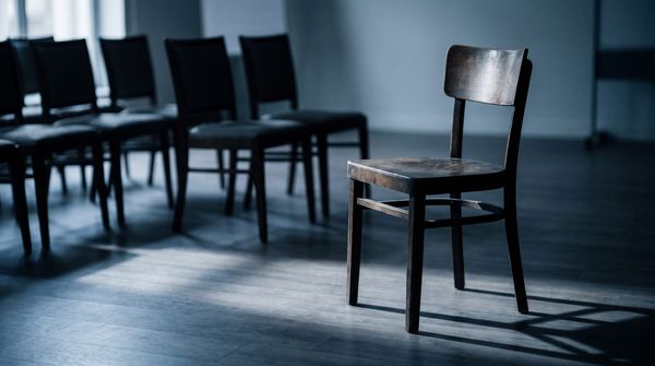 A single empty wooden chair kept open at the end of a row, in soft window light