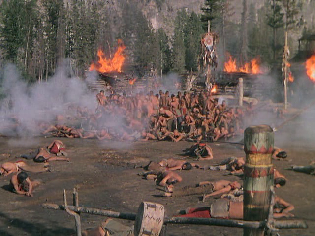 A group of Native Americans is huddled together. Around them lay the bodies of their compatriots, and wooden houses engulfed in flames.