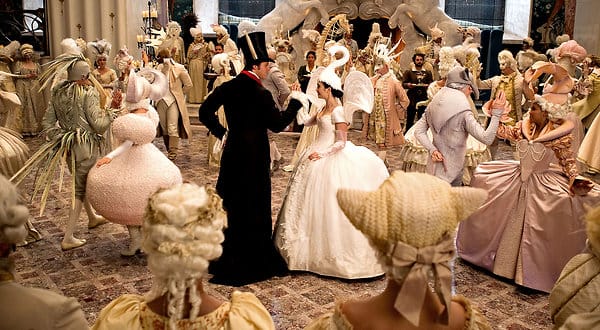 Amid a ballroom dance whose participants are dressed in animal-themed outfits, a man in a black cloak, top hat, and white rabbit ears, takes the hand of a woman in a white dress with outstretched swan wings from the back and a headpiece with an arched swan neck and head.