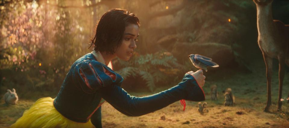 Snow White, but live-action, looking at a blue bird on her hand with concern. Around her are a forest and animals, none of which are real.