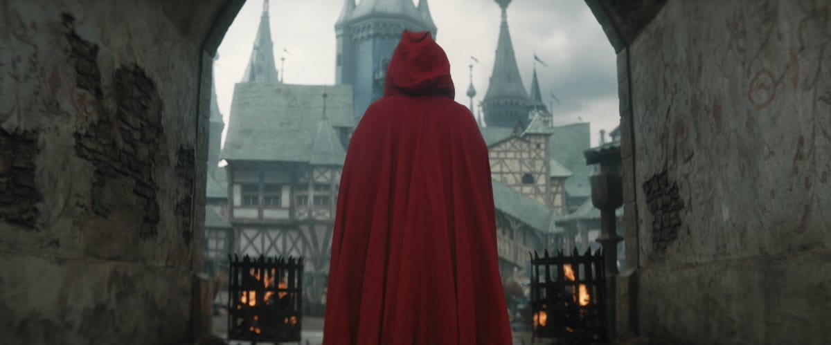 A figure draped in a red cloak steps through an archway toward a desolate, desaturated medieval town and castle.
