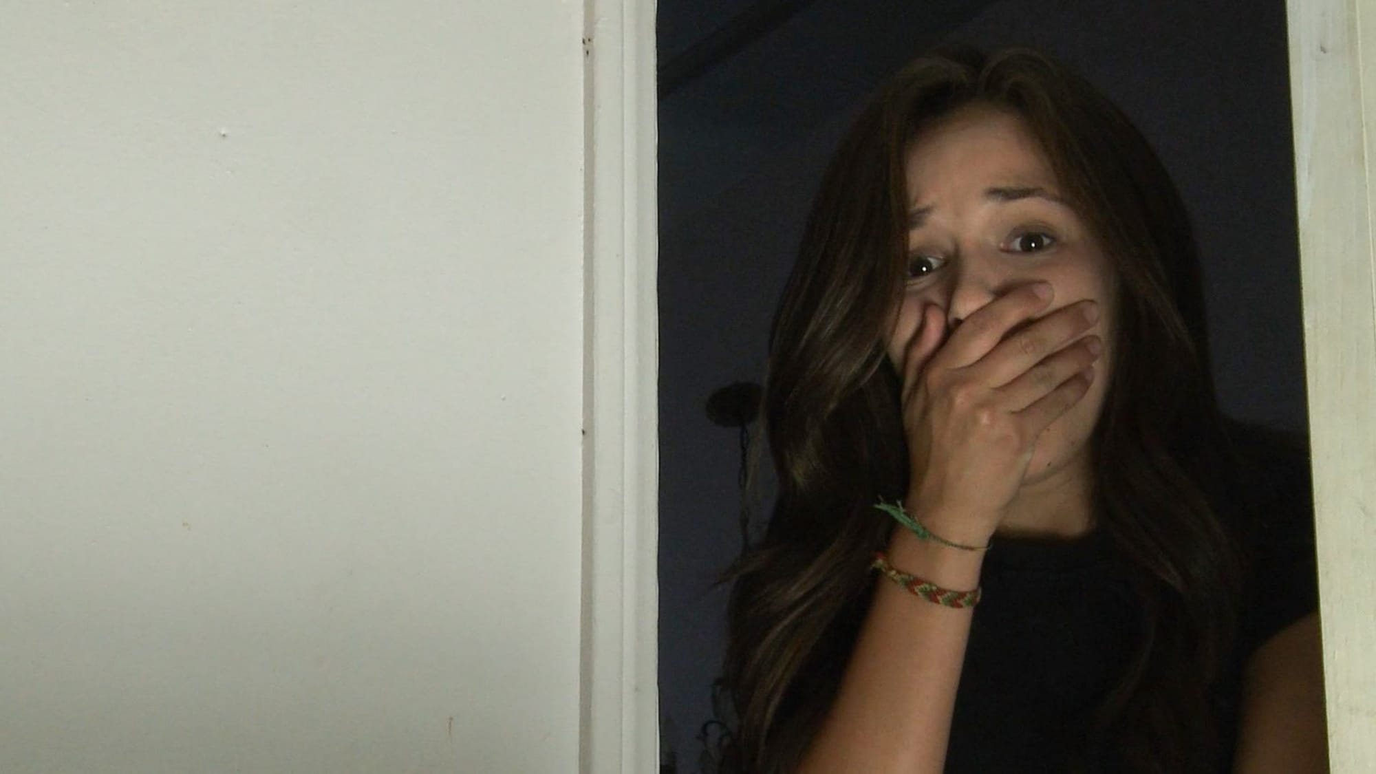 A young woman standing in a doorway puts her hand to her mouth in shock. She is all the way on the right side of the frame, with completely negative empty space on the left, and, like, come on.