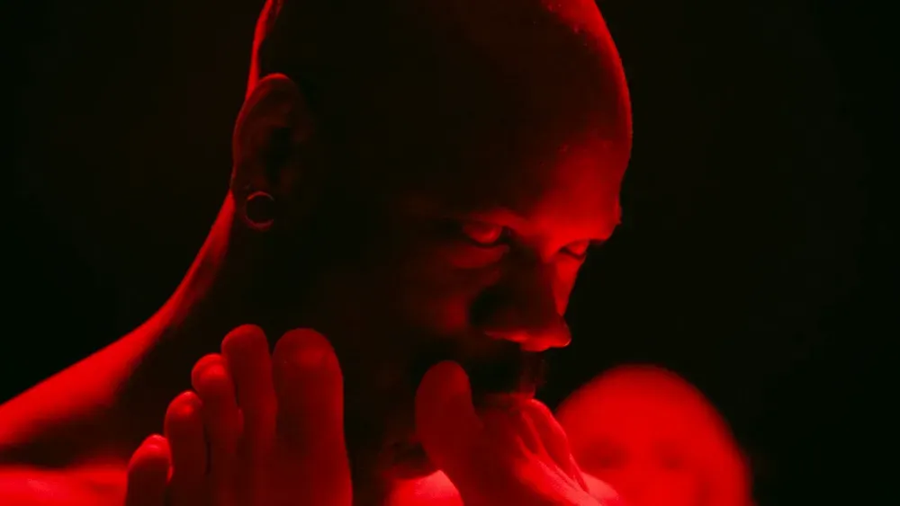 In red light, a bald-shaved men sucks the toes of a couple feet, his eyes solid white as if he were an alien.