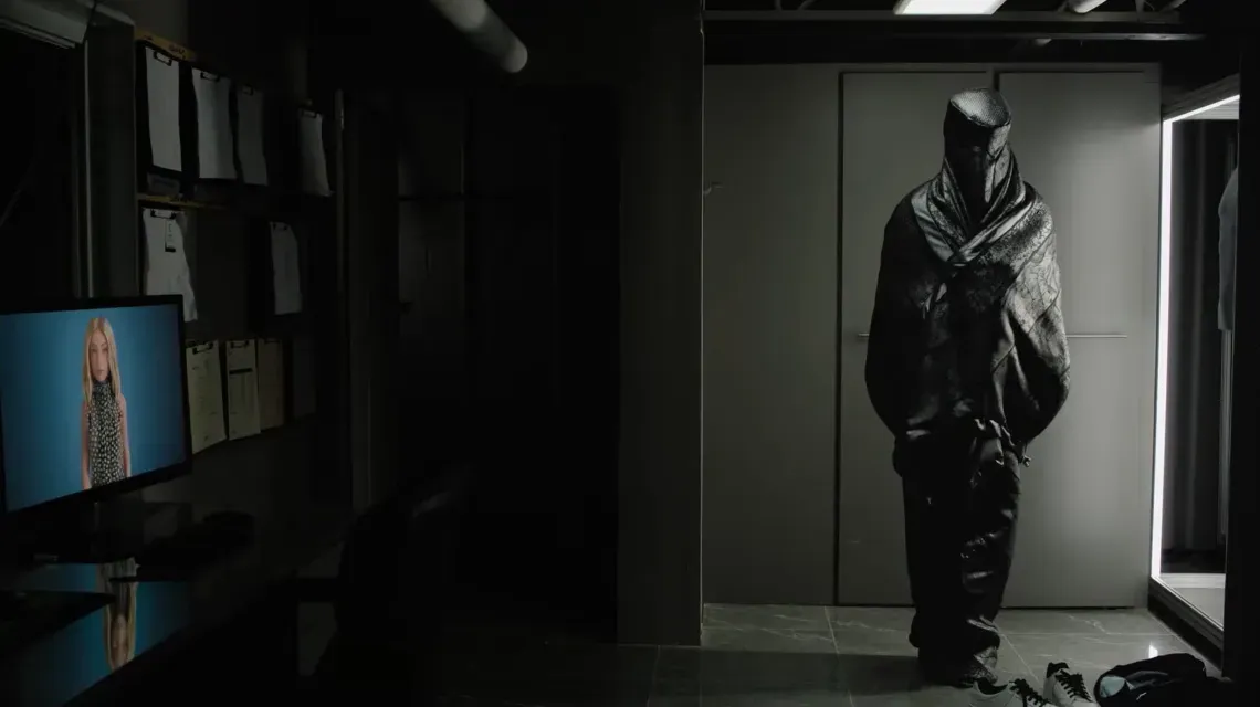 An office lit only LED light coming from a closet. At left, a computer monitor with CG female avatar. At right, in the light, figure wrapped in black shroud.