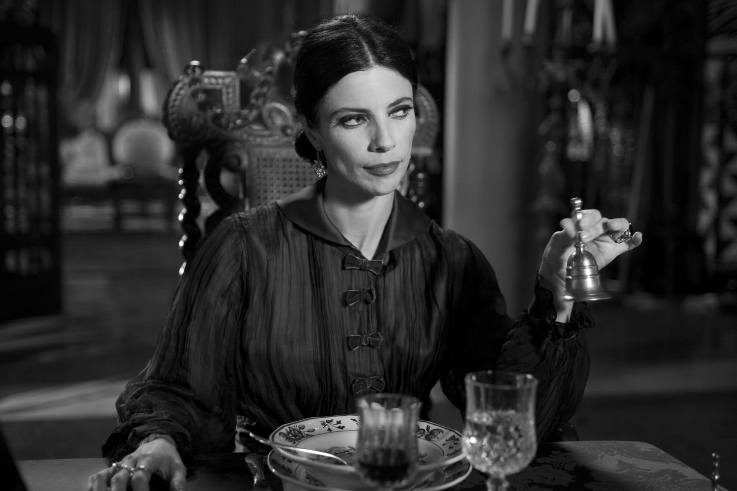 In black-and-white, a woman with black hair and blouse sits at the end of a dinner table and rings a sinister bell.