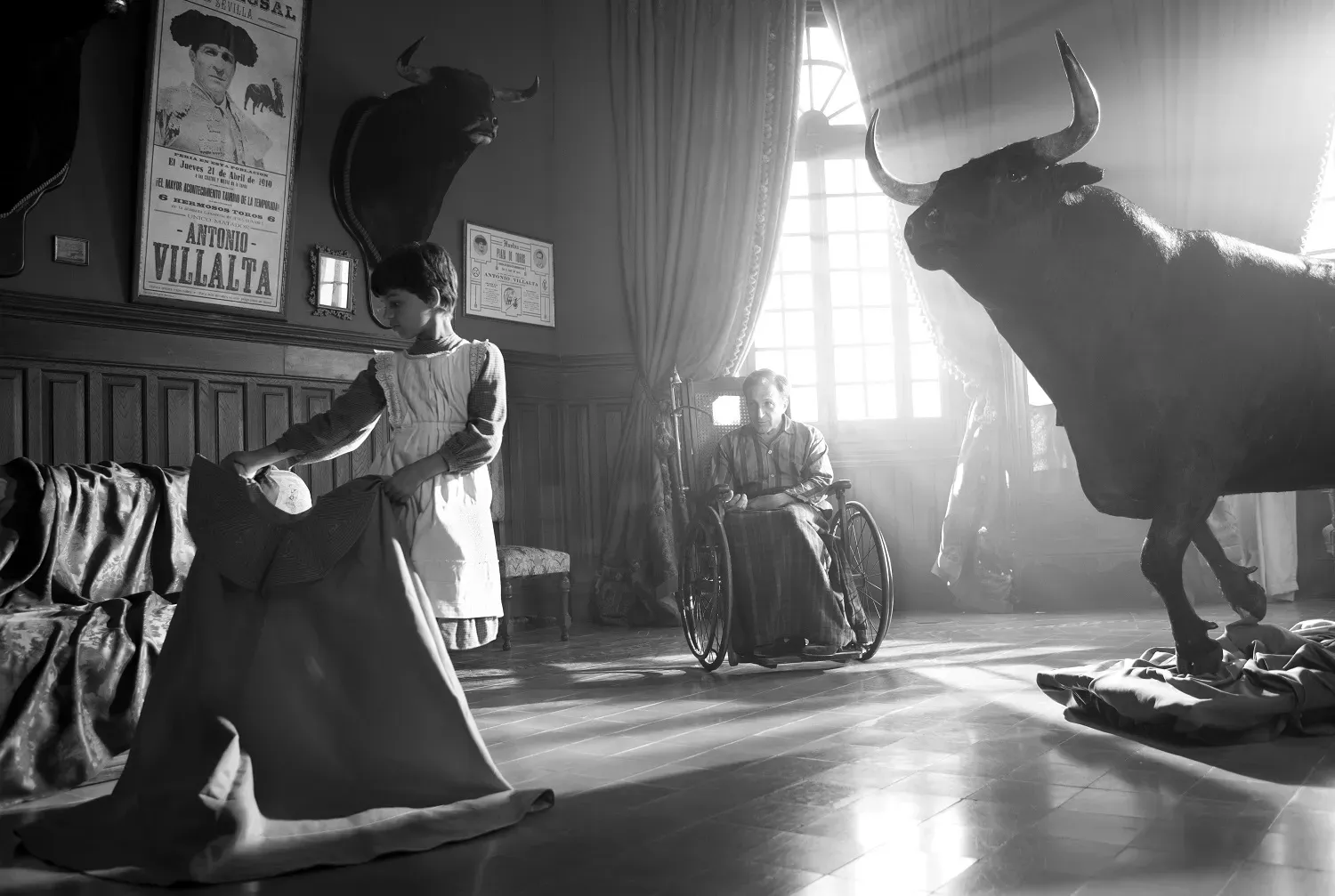 A young girl practices waving a matador's cloak. Behind her, a taxidermied bull. Between them, further back, an older man in a wheelchair. On the wall behind them is a poster of him in his prime, Antonio Villalta. A mounted bull head hangs next to it.