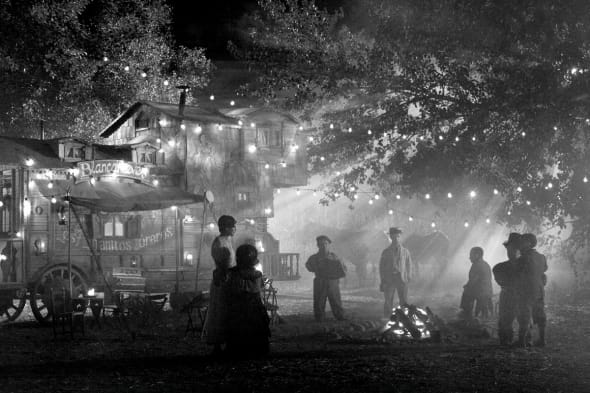 In black-and-white, several dwarfs and a young woman stand around a fire. Surrounding them are trees, through which god-rays of light stream through the mist, as well as a carnivalesque house strucutre.
