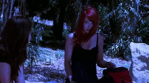 A young redheaded woman in a forest makes a vaguely incredulous gesture toward a brunette woman. Everything is filtered blue, and certain bright elements are too bright.