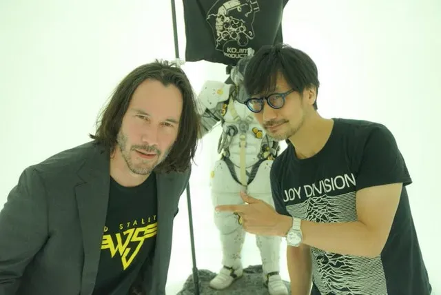 HIdeo Kojima, in a Joy Division shirt, points a finger left to Keanu Reeves, who smiles bemusedly, They stand in fron of an an astronaut-looking statue bearing a black flag with the Kojima Productions logo.