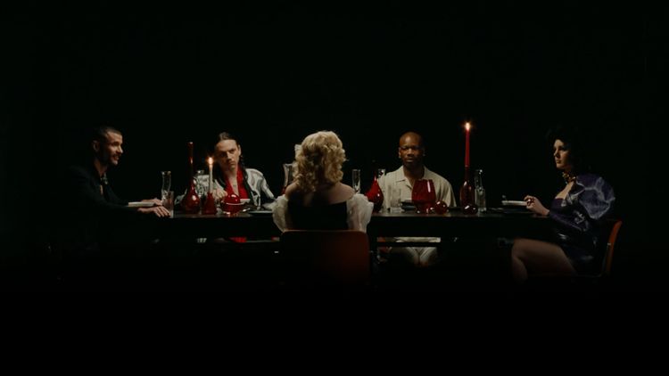 Lit by candles in an otherwise pitch-dark space, five people sit at a sleek dining table lit by candlelight.