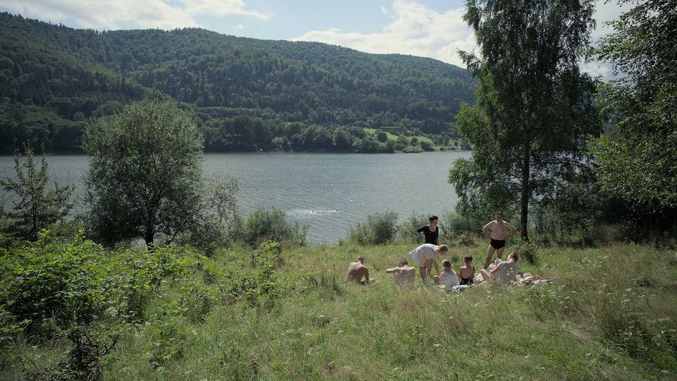 A screenshot from The Zone of Interest. A Nazi family picnics in the grass beside a river.