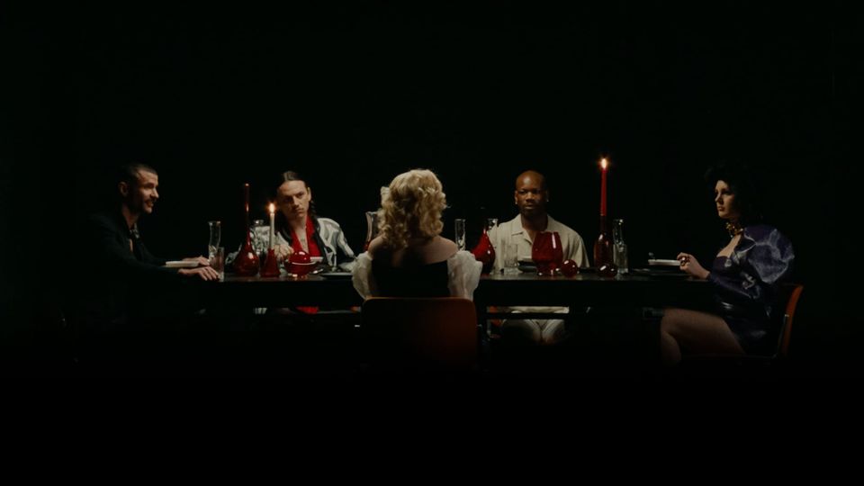 Lit by candles in an otherwise pitch-dark space, five people sit at a sleek dining table lit by candlelight.
