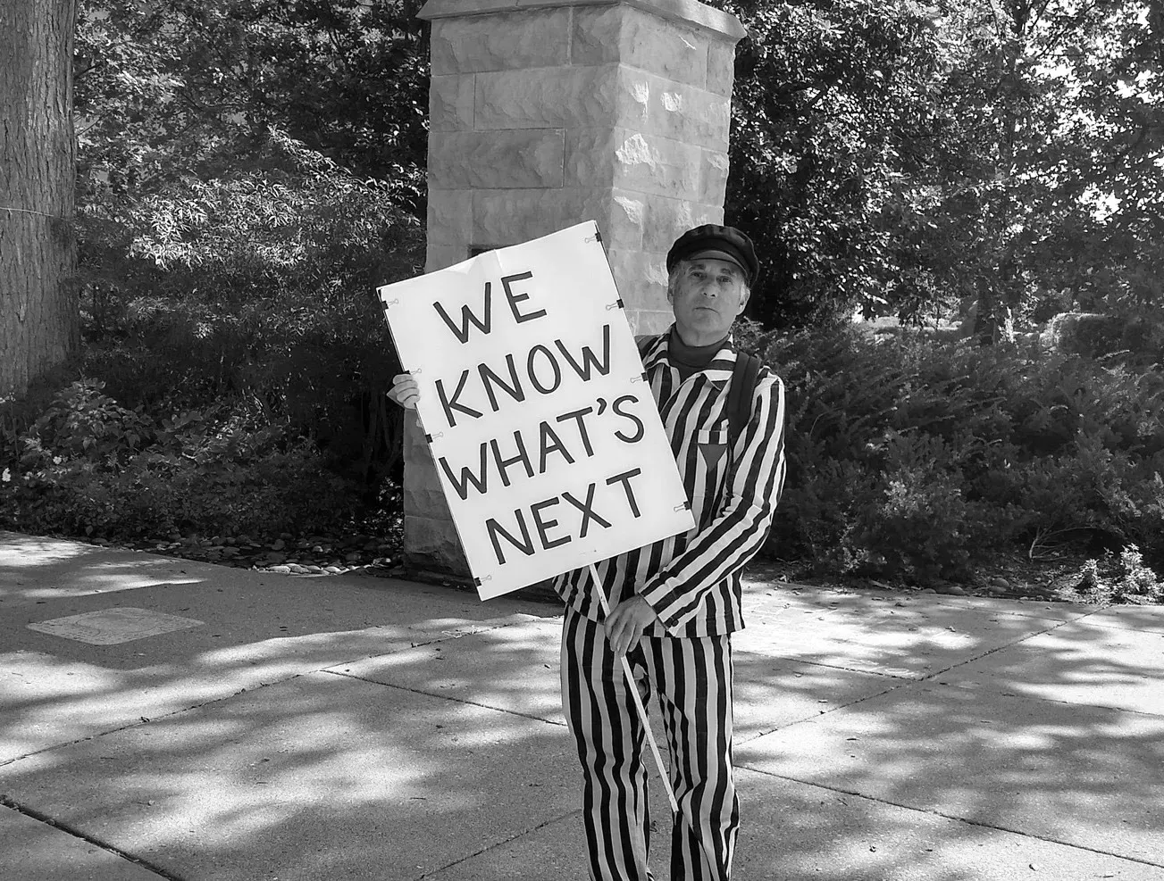 A black and white photo of a man in a striped prison suit with a triangle on the front holding a sign that says "We know what's next"