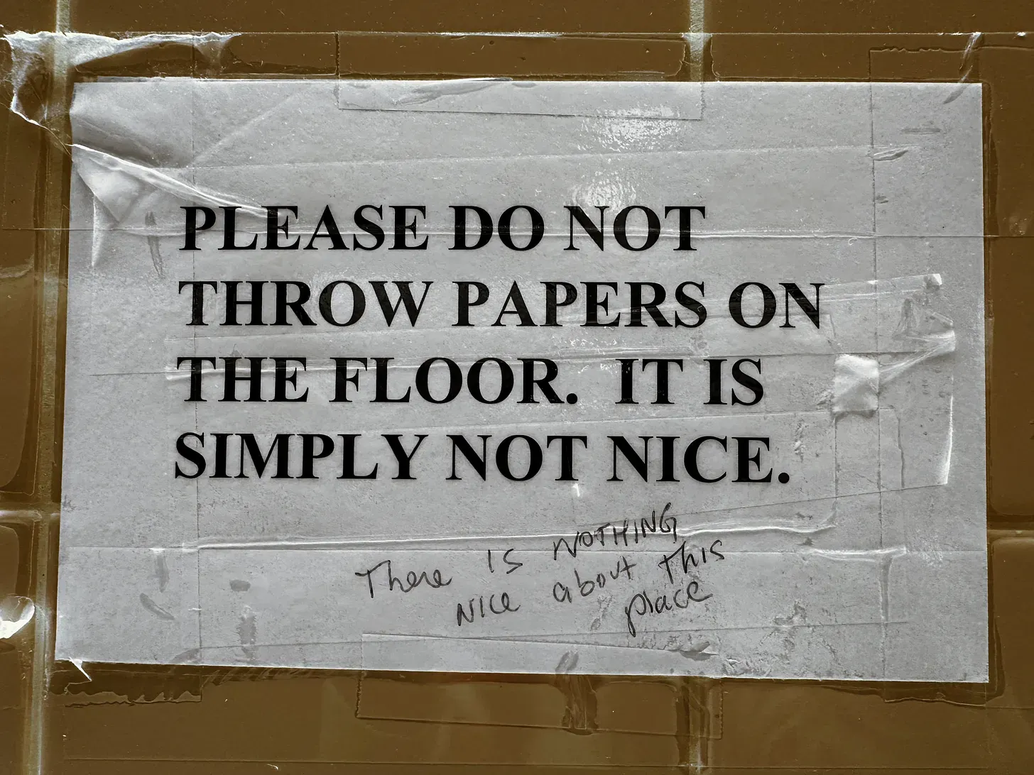 A printed signed taped to the wall says "Please do not throw papers on the floor. It is simply not nice" Scrawled in pen "There is nothing nice about this place"