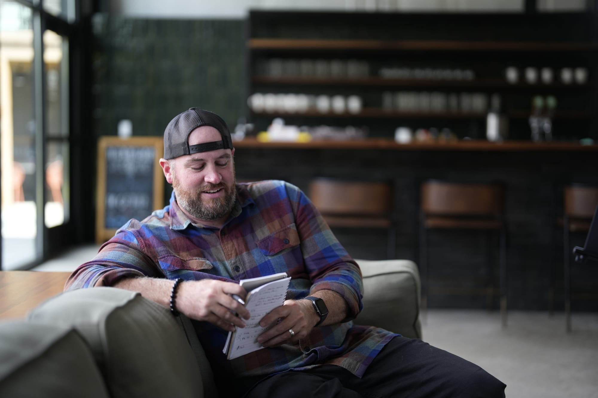 Thomas, sitting on a couch in a wework looking cafe, grinning at the notebook in his hands like it has a stupid dad joke written on it.