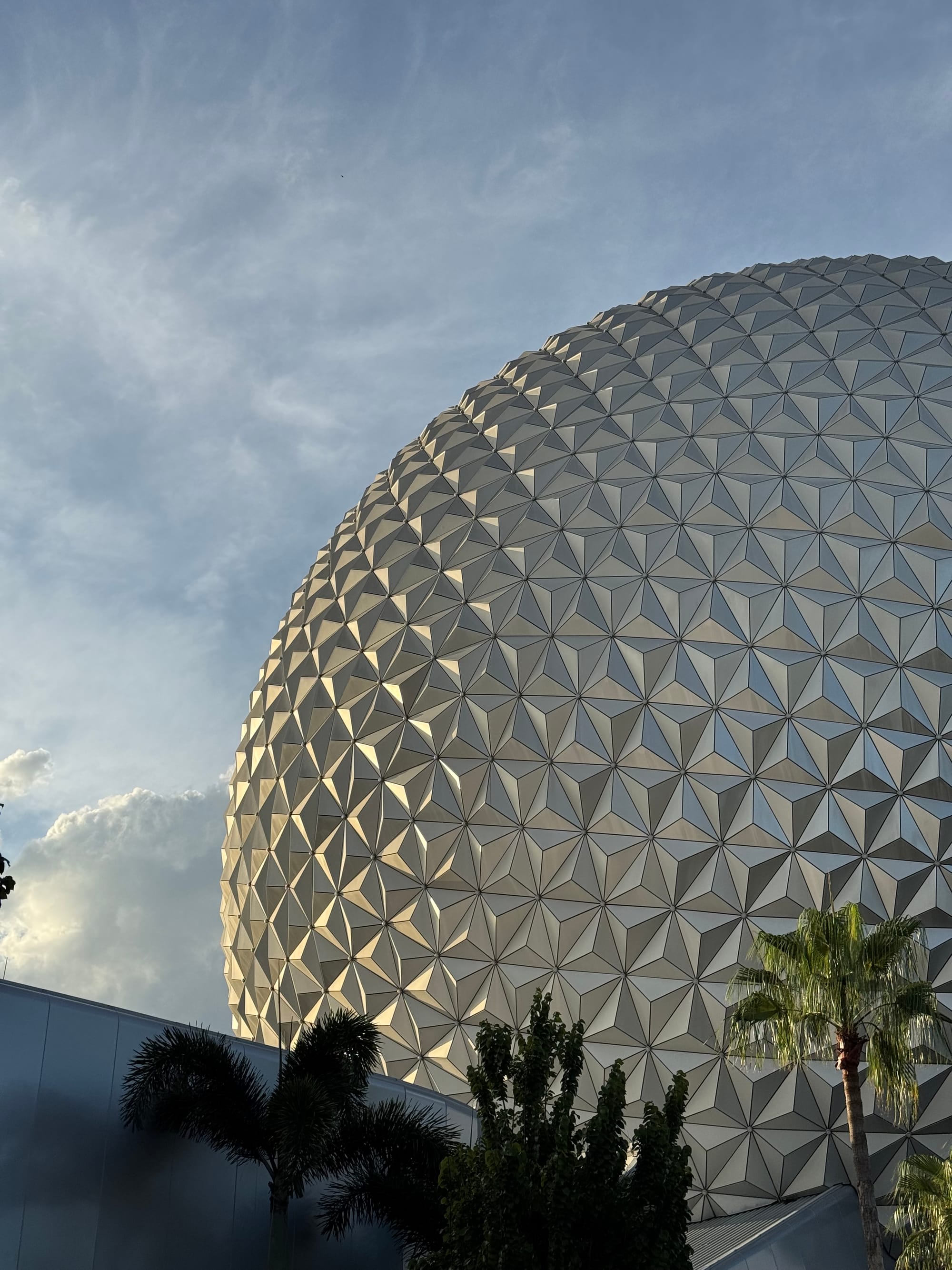 Spaceship Earth at Epcot, shown from a low angle, with its silver geometric panels catching soft evening light, framed by palm trees and nearby buildings under a lightly clouded sky.
