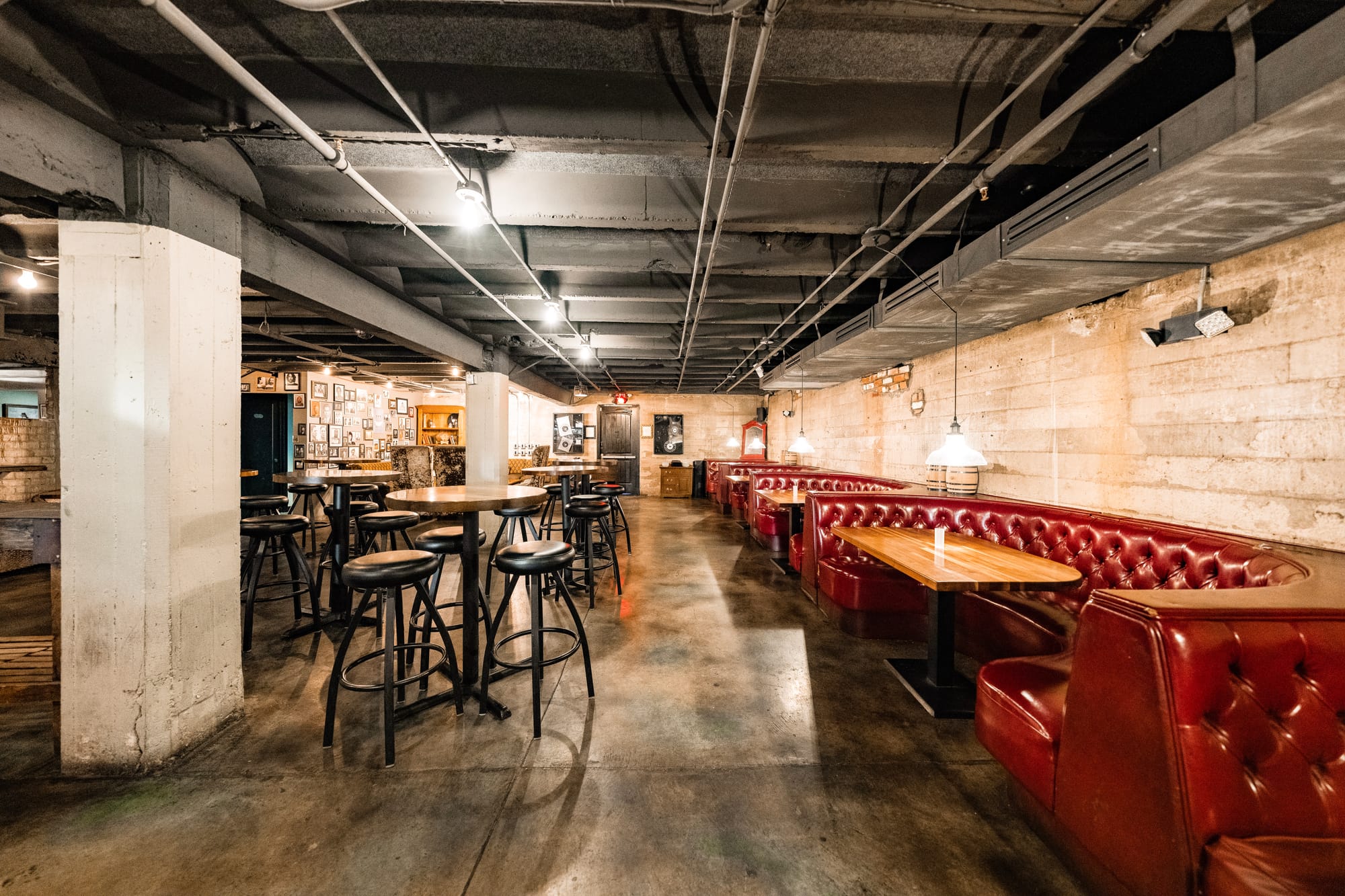 A wide and inviting speakeasy interior with large crescent shaped red booths, exposed brick walls, and high-top seating. In the back, a wall is decorated with framed vintage celebrity photos.