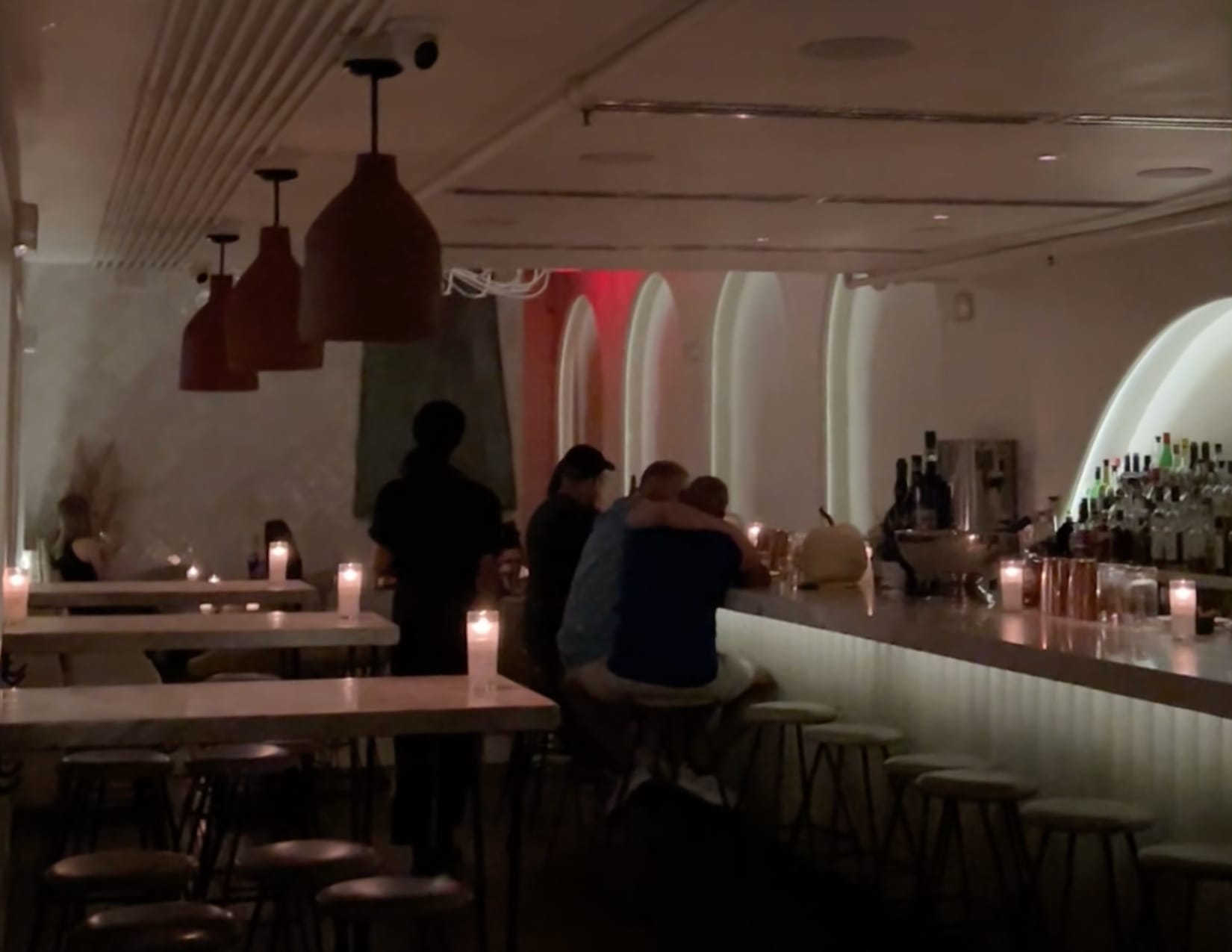 A classy dimly-lit room with white ceilings, candle-lit wooden tables, bar stools and a glowing arch of light behind the bar. Speakeasy patrons embrace and enjoy a drink at the bar top.