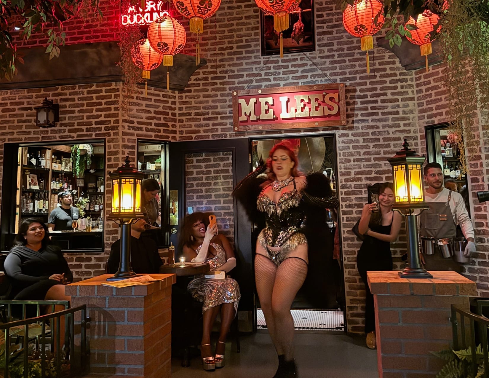 A burlesque dancer poses outside the speakeasy, framed by brick walls, a sign above the door that reads M.E. LEES and vibrant red lanterns. Guests enjoy the lively atmosphere, surrounded by lush greenery.