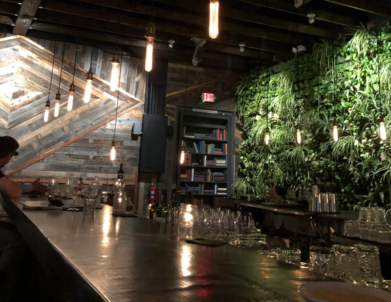 Dimly lit room featuring a lush greenery-filled wall behind the bar, with another wall adorned in intricate wooden panels. A hidden bookshelf door serves as the exit, and lights hang from the ceiling, adding to the cozy atmosphere of this speakeasy.