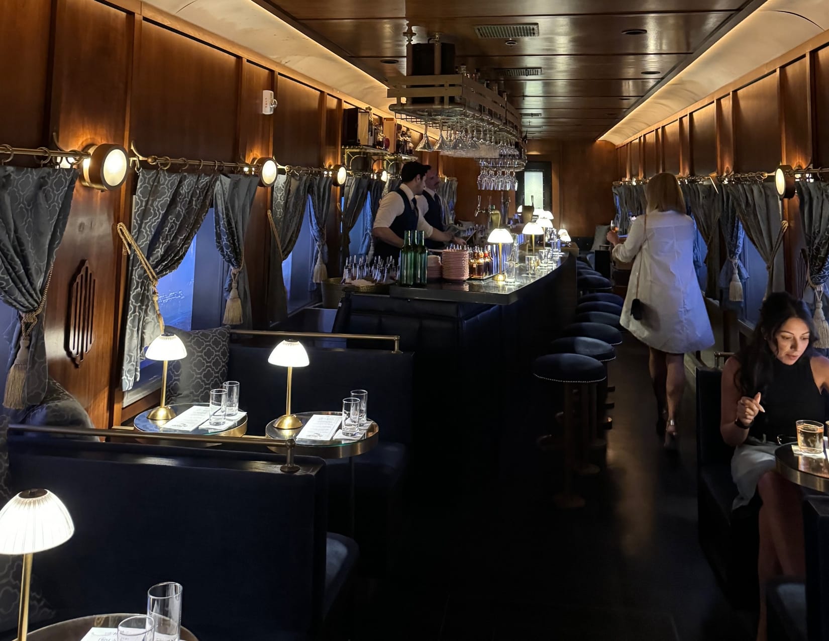 A speakeasy designed as a replica of a 1927 Pullman train car with art deco decor, including gold lamps on each small table, blue velvet seating, and wine glasses hanging above a small bar. A moving skyline is shown on the windows and the servers are dressed as conductors. 