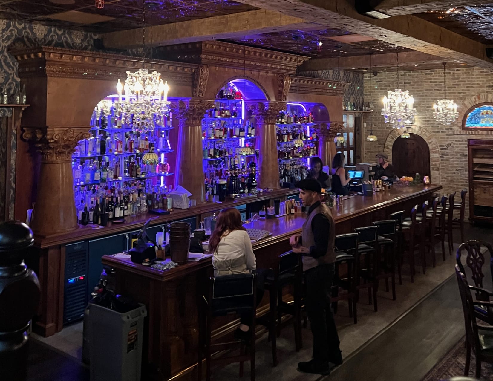 Beautiful columns surround the purple glowing back bar of this speakeasy. Brick walls, ornate wallpaper, chandeliers and a stained glass window add a warm and stunning ambiance.