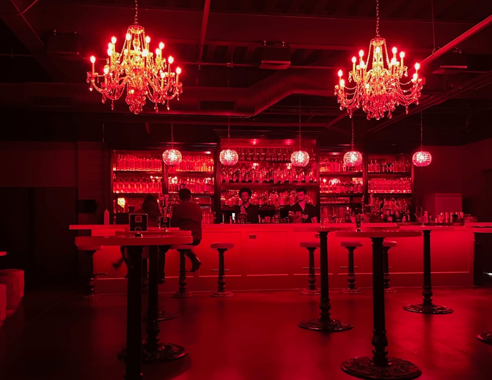 The Parlor Room speakeasy glows with red lighting, crystal chandeliers, bar seating, and illuminated shelves of glassware. Bartenders chat up patrons at the bar.