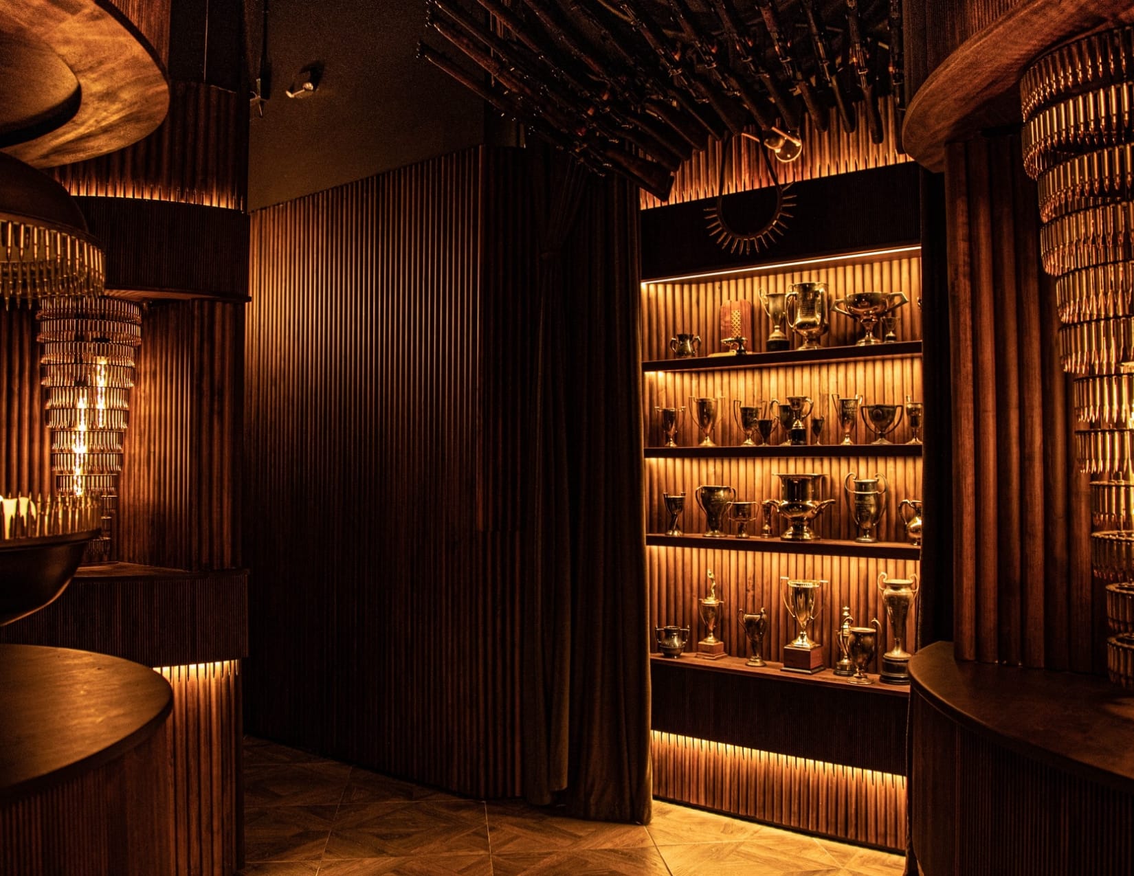 Art Deco inspired speakeasy interior with amber illuminated flute walls, warm woof flooring, and a bookcase filled with golden trophies.