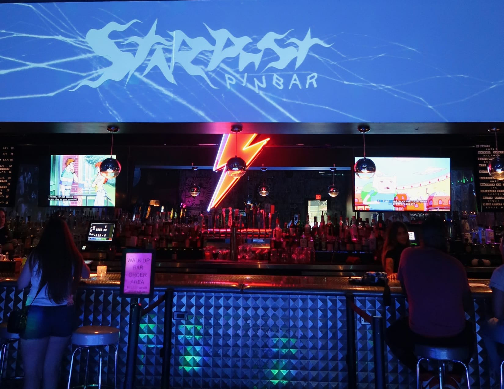 Dimly lit room with a glowing neon lightning bolt above the bar. 'Stardust Pinbar' is in intricate font above the bar, and a sign indicates the 'Walk up bar order area.' Guests enjoy cocktails while watching TV, creating a relaxed atmosphere for this speakeasy.