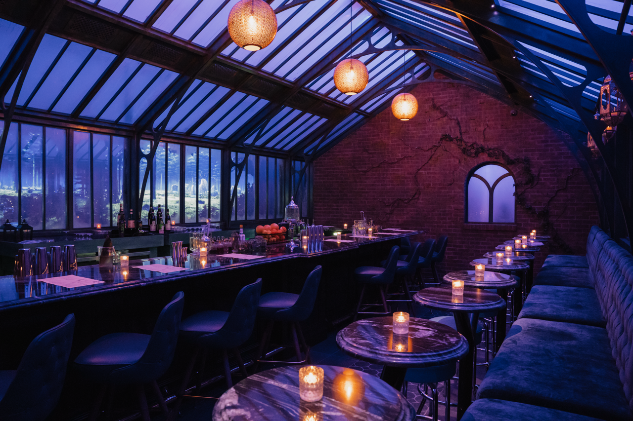 A whimsical speakeasy with a glowing purple aura, velvet seats at round tables with candles, and a brick wall with vines growing across. The bar stretches along one side with a long counter and seats in front, while animated fairy scenes light up the walls and ceiling.