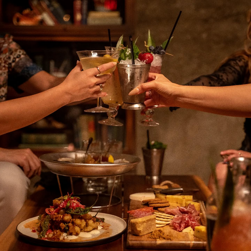 Friends clink cocktails over a table filled with charcuterie in a dimly lit bar.