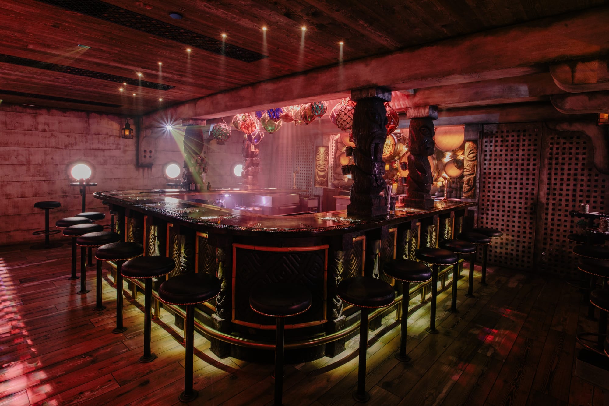 A large bar is surrounded by bar stools and glowing lights in a dimly lit recreation of a clipper ship hull. The speakeasy is decorated with unique tiki decor, dark wooden flooring and walls displaying porthole windows.