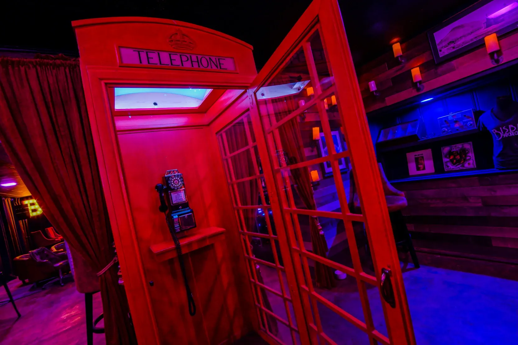 A vibrant scene of this speakeasy featuring a red telephone booth with the door open, red curtains framing the entryway to another room surrounded by a neon pink and blue glow. The wall wooden next to the booth is lined with orange accent lights and photos.