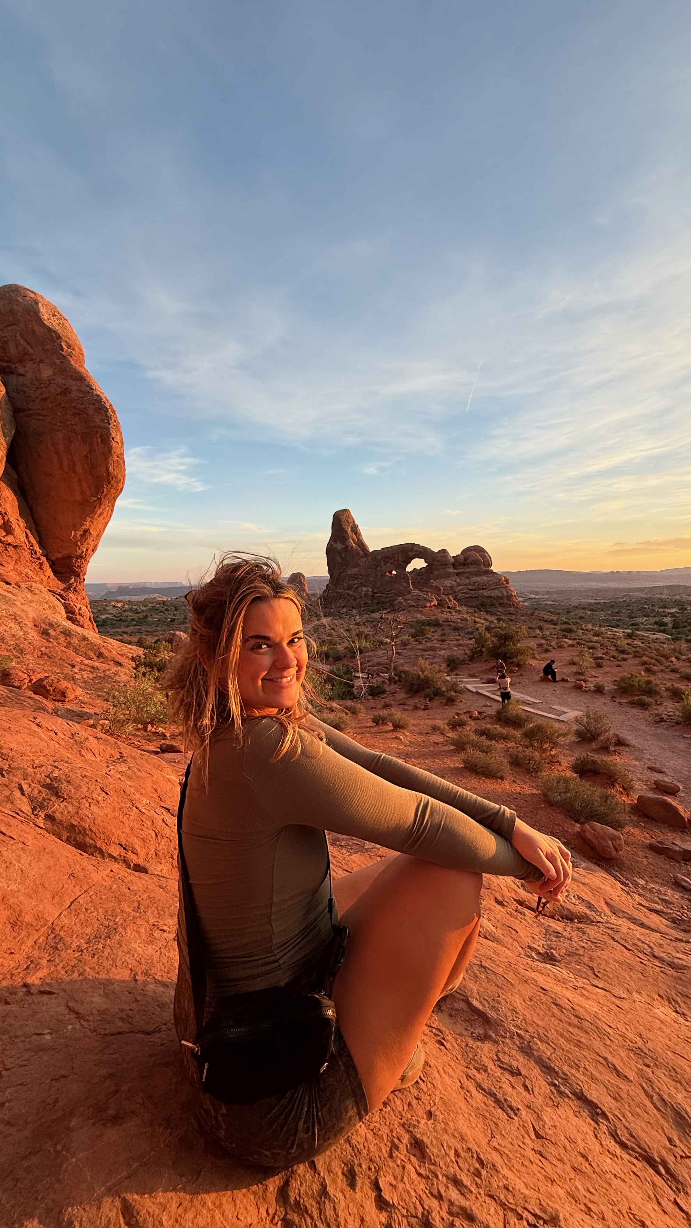 woman sitting on sandstone rocks during sunset with Turret Arch in the background at Arches National Park in Moab, Utah