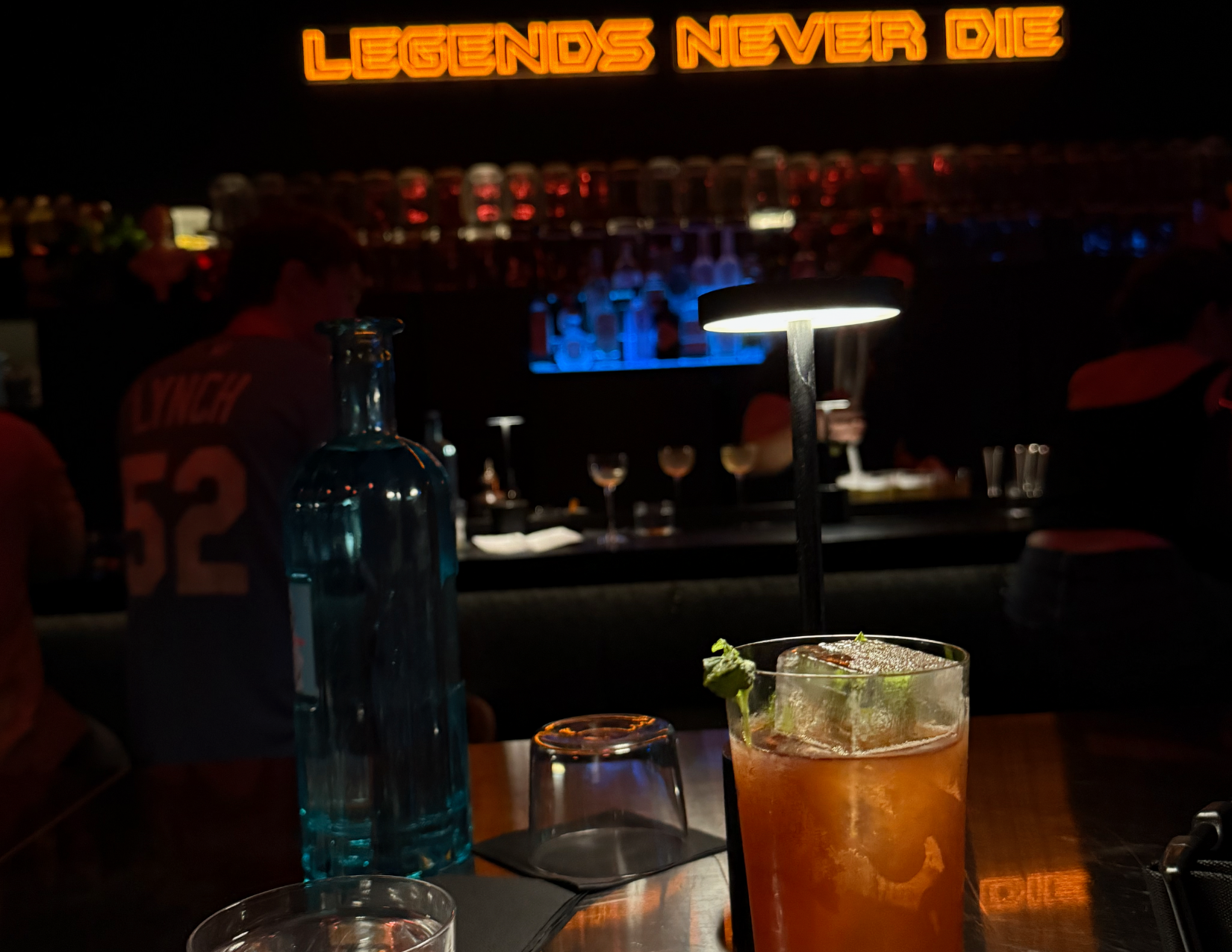 A craft cocktail sits on a table beneath a glowing orange 'Legends Never Die' neon sign. 