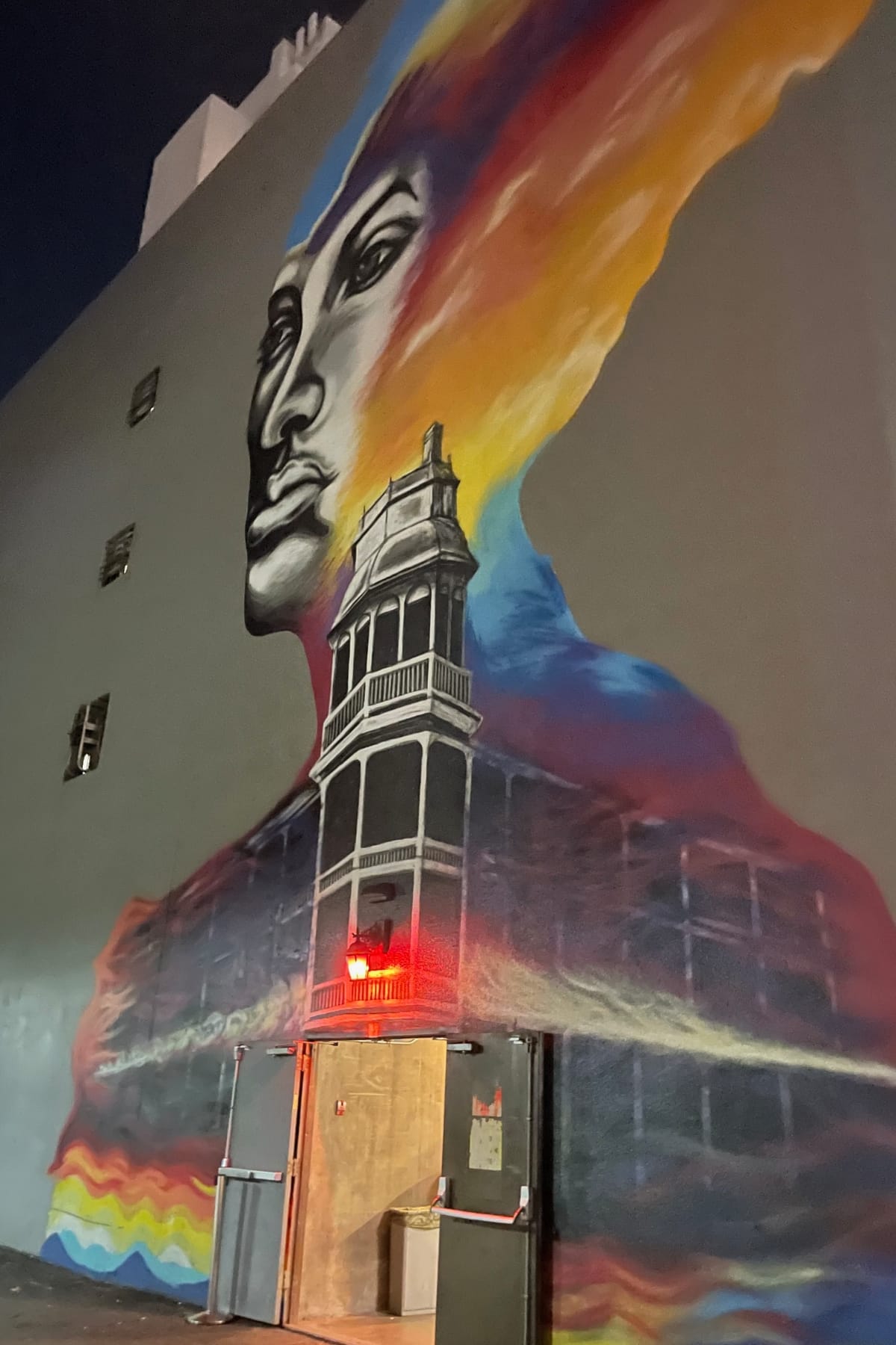 Large colorful mural outside Melinda's alley in Downtown Phoenix featuring a woman's face and historic building, with the hidden speakeasy entrance illuminated by a red light.