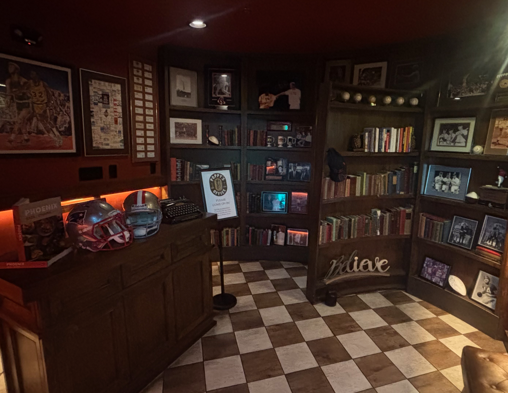 A cozy speakeasy with floor-to-ceiling bookshelves, vintage memorabilia, framed photographs, and checkered flooring.