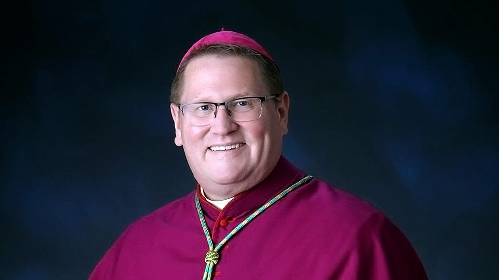 Bishop in clerical attire smiles against a dark background, wearing magenta robes, square glasses and a zucchetto.