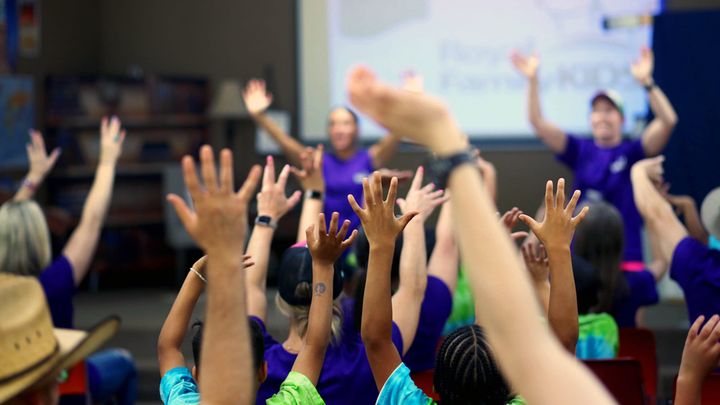 A group of individuals wearing purple shirts enthusiastically raising their hands in a display of excitement or participation.