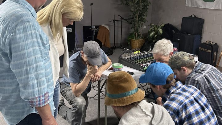 Several people dressed in casual clothing kneel, stand, and sit with heads bowed.