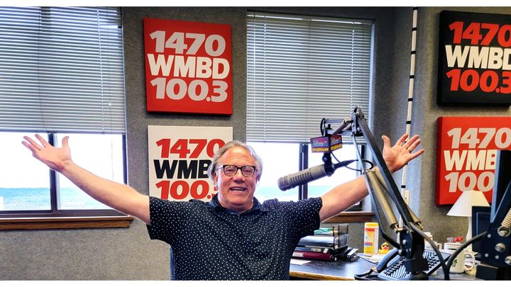 Radio host with arms outstretched in studio with WMBD 1470 signage. Celebrating on-air.