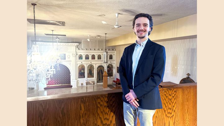 A young man in a blue blazer stands smiling in the balcony of a Greek Orthodox church.