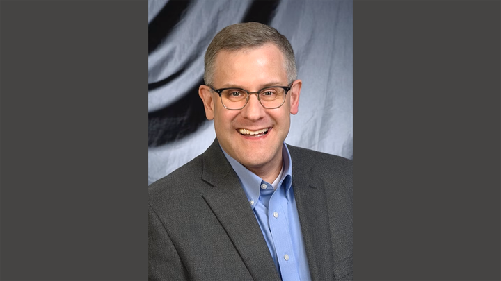 A smiling man in a blue shirt and gray blazer looks at the camera.